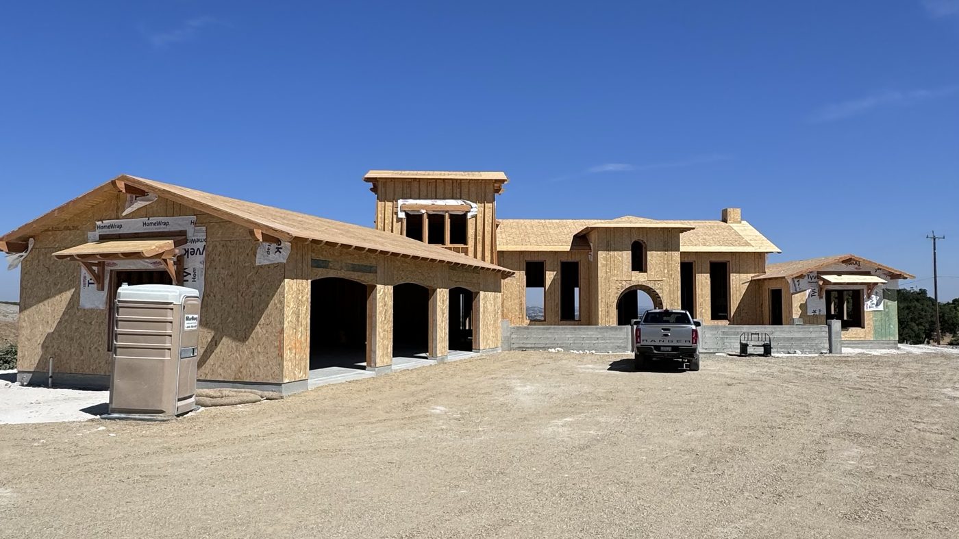 A large house in framing, sheathed in plywood, on a graded dirt lot under a blue sky.