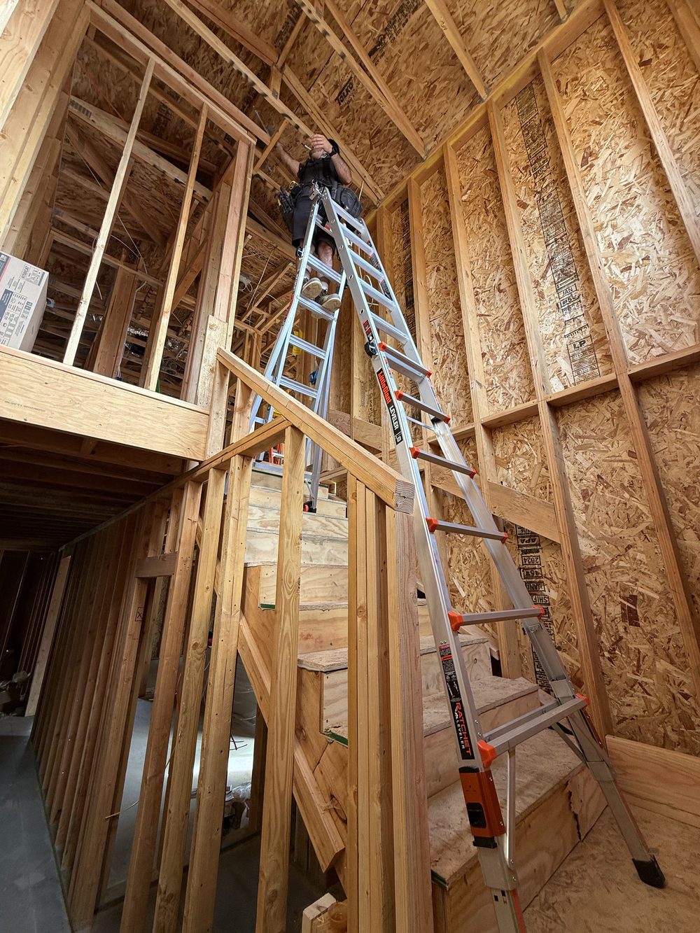 An electrician in a tool belt at the top of a tall ladder, working on the framing of a high ceiling.